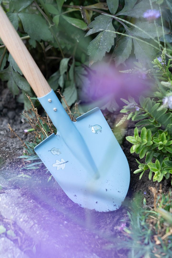 Blaue Kinder-Gartenschaufel mit Holzstiel beim Buddeln in einem Blumenbeet. Robustes Gartenspielzeug für Kinder zum Graben, Pflanzen und Entdecken im Garten.