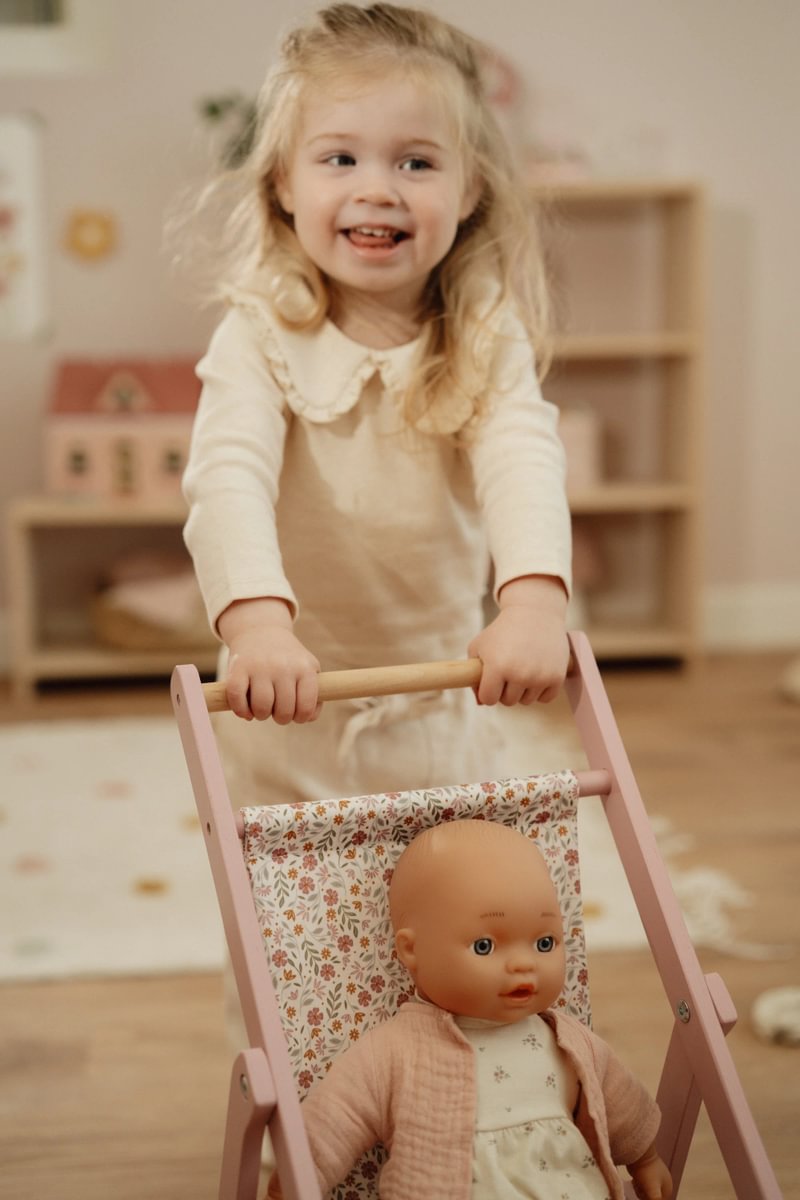Kind schiebt einen Puppenbuggy aus Holz mit Babypuppe im Kinderzimmer. Beliebtes Geschenk für Kinder, das Rollenspiele, Kreativität und fantasievolles Spielen fördert.